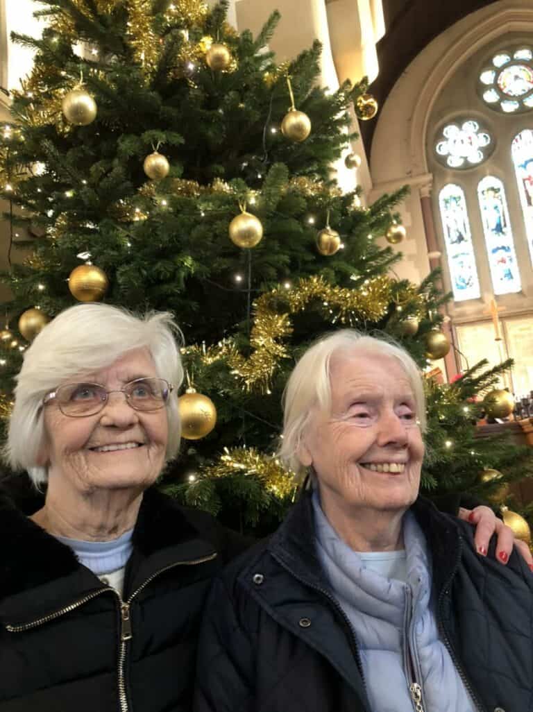 Two older women stand in front of a Christmas tree in March with gold baubles.
