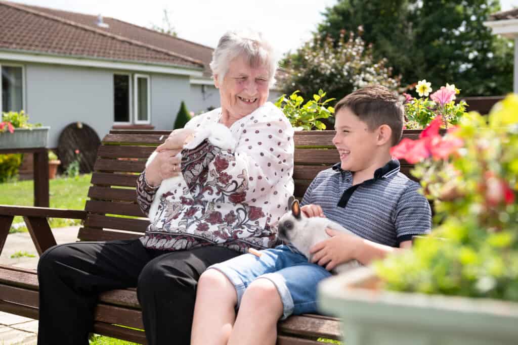 Boy sits on bench in garden of care home with older woman and they stroke a bunny rabbit