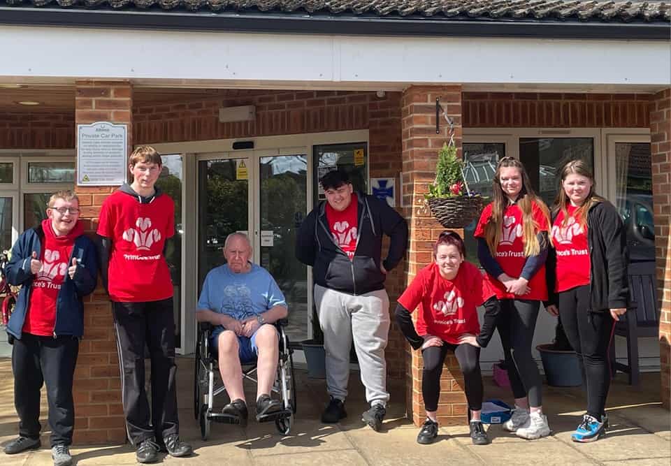 Group of princes trust volunteers wearing red tee shirts outside care home with older man in wheelchair